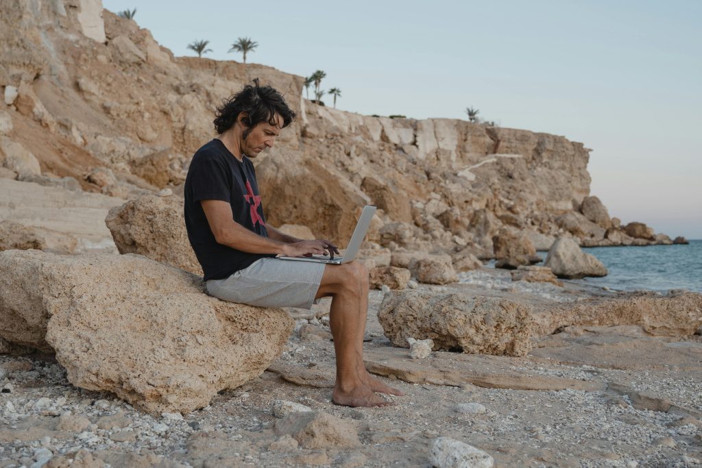 Man sits on rocks with laptop by the beach, enjoying remote work under a serene sunset.